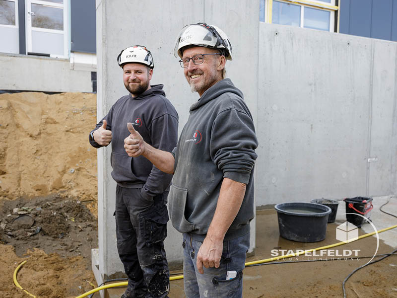 Zwei Mitarbeiter der Stapelfeldt Bauunternehmung vor Betonwand im Hallenbau Norderstedt, Daumen hoch auf der Baustelle.